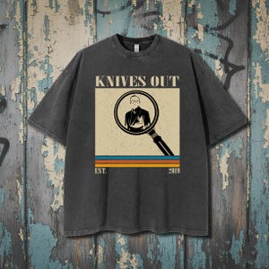 May include: Dark gray t-shirt featuring the text "KNIVES OUT" above a graphic of a person in a magnifying glass. Below the graphic are horizontal stripes in orange, blue, and yellow, with "EST. 2019" below the stripes.