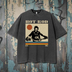 May include: Dark grey t-shirt with a vintage graphic. The graphic features the text "HOT ROD" above an illustration of a motorcyclist. Below the illustration are the words "EST. 2007" and horizontal stripes in orange, blue, and yellow.