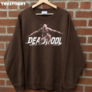 May include: A brown sweatshirt with a graphic of Deadpool kneeling with swords. The word "DEADPOOL" is printed in white, distressed lettering. The word "SWEATSHIRT" is printed at the top of the image.