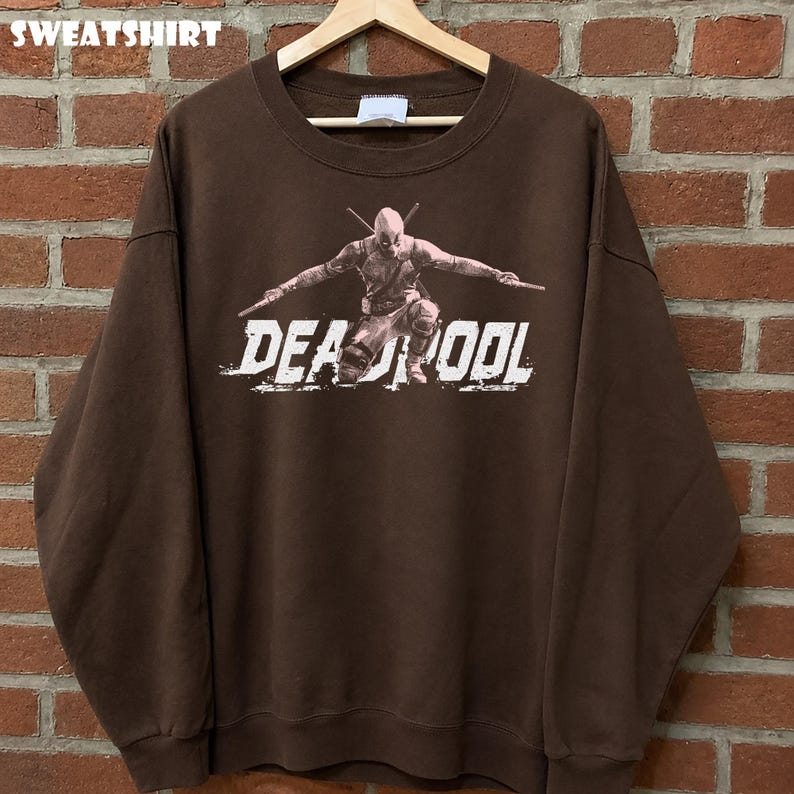 May include: A brown sweatshirt with a graphic of Deadpool kneeling with swords. The word "DEADPOOL" is printed in white, distressed lettering. The word "SWEATSHIRT" is printed at the top of the image.