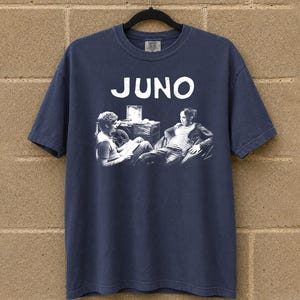 Peut inclure: T-shirt bleu marine avec le mot "JUNO" en blanc. En dessous, un dessin en noir et blanc de deux personnes assises. Le t-shirt est suspendu à un cintre noir devant un mur de briques.