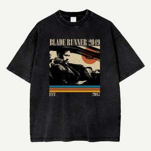 May include: Black t-shirt featuring a graphic from the movie "Blade Runner 2049." The design includes the film's title, a retro image of a man in a car, and the year "2017" below colorful stripes. The shirt has a vintage, washed look.