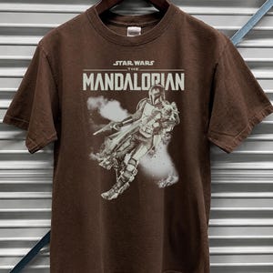 May include: Dark chocolate brown t-shirt with a graphic of The Mandalorian from Star Wars. The text "STAR WARS THE MANDALORIAN" is printed across the chest. The shirt is short-sleeved and is displayed on a wooden hanger.