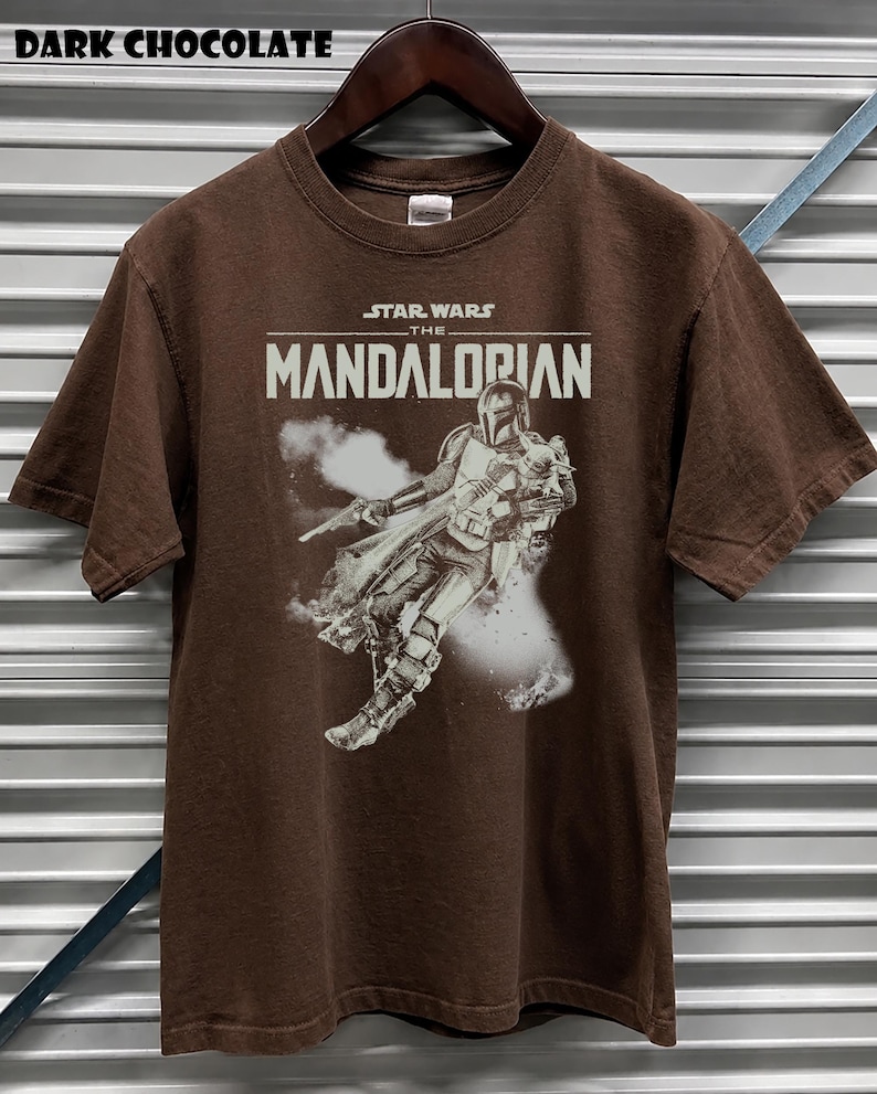 May include: Dark chocolate brown t-shirt with a graphic of The Mandalorian from Star Wars. The text "STAR WARS THE MANDALORIAN" is printed across the chest. The shirt is short-sleeved and is displayed on a wooden hanger.
