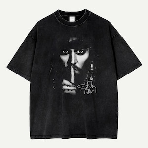 Puede incluir: Camiseta negra con un estampado en blanco y negro de una persona con un dedo sobre los labios. La imagen tiene una firma blanca. La camiseta tiene mangas cortas y cuello redondo.