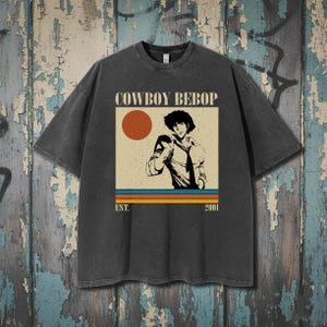 Puede incluir: Camiseta gris oscuro con un diseño de la serie de anime Cowboy Bebop. El diseño incluye el texto "COWBOY BEBOP" sobre una ilustración de un personaje, un sol rojo y rayas horizontales. El texto "EST. 2001" está debajo.