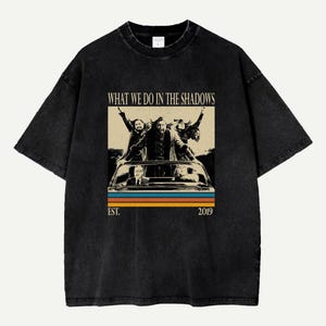 Könnte beinhalten: Schwarzes T-Shirt mit einem Grafikdesign aus der Fernsehserie "What We Do in the Shadows". Das Design zeigt die Charaktere in einem Auto, mit dem Text "WHAT WE DO IN THE SHADOWS" und "EST. 2019" im Vintage-Stil.