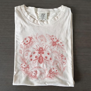 May include: A cream-coloured short-sleeve t-shirt featuring a red ink graphic of multiple ladybirds surrounded by botanical elements. The interior neck label displays the text Comfort Colors and size M. The design is centred on the front of the shirt.