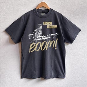 May include: Dark gray t-shirt featuring a black and white image of a person at a desk, with the words "tick, tick..." above and "BOOM!" below in a distressed font. The shirt is hanging on a wooden hanger.
