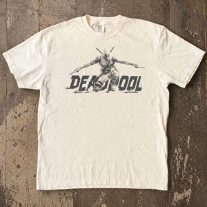 May include: Cream-coloured t-shirt featuring a graphic of Deadpool kneeling with weapons drawn. The word "DEADPOOL" is printed in a distressed font below the character. The shirt has short sleeves and a crew neck.