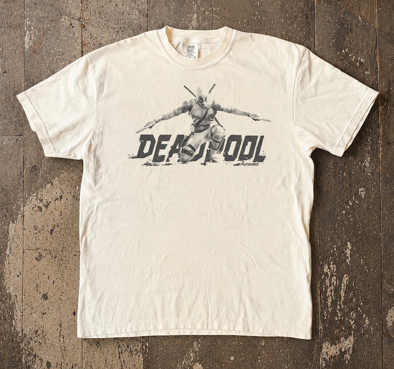 May include: Cream-coloured t-shirt featuring a graphic of Deadpool kneeling with weapons drawn. The word "DEADPOOL" is printed in a distressed font below the character. The shirt has short sleeves and a crew neck.