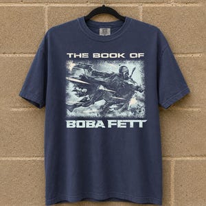 May include: Navy blue t-shirt featuring a graphic from "The Book of Boba Fett." The design shows Boba Fett in action with the text "THE BOOK OF" above and "BOBA FETT" below the image. The shirt is hanging on a black hanger.