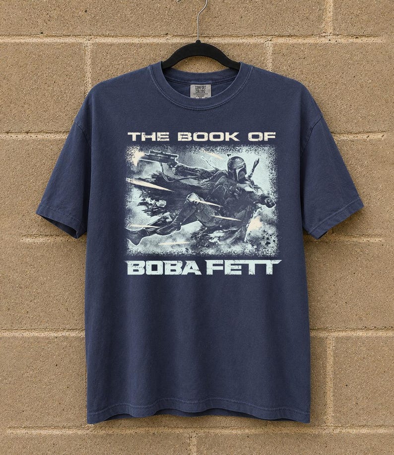 May include: Navy blue t-shirt featuring a graphic from "The Book of Boba Fett." The design shows Boba Fett in action with the text "THE BOOK OF" above and "BOBA FETT" below the image. The shirt is hanging on a black hanger.