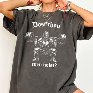 May include: A charcoal gray short-sleeve t-shirt featuring a white graphic of a knight in full armor performing a barbell squat. The text above the knight reads "Dost thou" and the text below reads "even hoist?" in a gothic-style font.