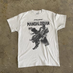 May include: White t-shirt with a black and grey graphic of The Mandalorian from Star Wars. The text "STAR WARS THE MANDALORIAN" is printed above the image. The shirt has a crew neck and short sleeves.