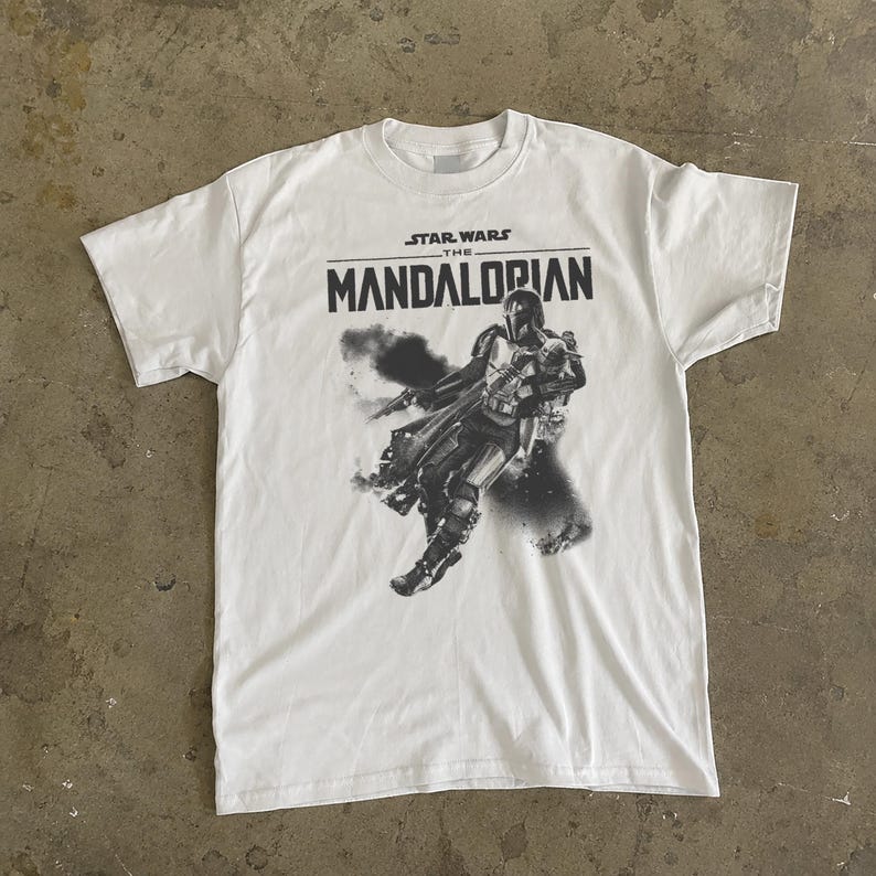 May include: White t-shirt with a black and grey graphic of The Mandalorian from Star Wars. The text "STAR WARS THE MANDALORIAN" is printed above the image. The shirt has a crew neck and short sleeves.