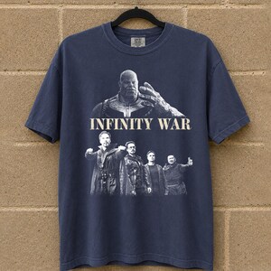 K&ouml;nnte beinhalten: Marineblaues T-Shirt mit einer Grafik aus dem Film "Infinity War". Das Design zeigt den Text "INFINITY WAR" &uuml;ber einer Darstellung von Thanos und anderen Charakteren. Das T-Shirt h&auml;ngt an einem schwarzen Kleiderb&uuml;gel.