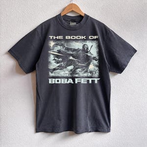 May include: Dark gray t-shirt featuring a graphic from "The Book of Boba Fett." The design shows Boba Fett in action with the text "THE BOOK OF" above and "BOBA FETT" below. The shirt is hanging on a wooden hanger.