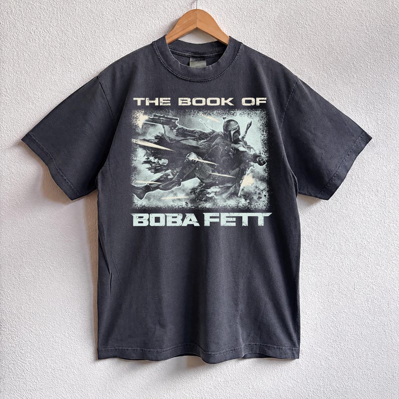 May include: Dark gray t-shirt featuring a graphic from "The Book of Boba Fett." The design shows Boba Fett in action with the text "THE BOOK OF" above and "BOBA FETT" below. The shirt is hanging on a wooden hanger.