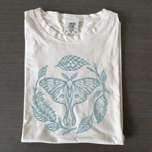 Op de afbeelding: Een crèmekleurig t-shirt met een blauwgroene grafische print van een mot, een rups, een cocon en bladeren in een cirkelvormig patroon. Het label in de nek bevat de tekst Comfort Colors en de maataanduiding M op het kledingstuk.