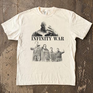 K&ouml;nnte beinhalten: Cremefarbenes T-Shirt mit einer Grafik von Thanos und anderen Charakteren aus dem Film "Infinity War". Der Text "INFINITY WAR" ist in fetten, schwarzen Buchstaben unter dem Thanos-Bild aufgedruckt. Das Shirt hat einen Rundhalsausschnitt und kurze &Auml;rmel.