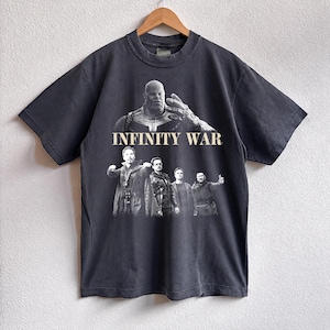 K&ouml;nnte beinhalten: Dunkelgraues T-Shirt mit einer Grafik aus dem Film "Infinity War". Das Design zeigt Charaktere und den Titel in Wei&szlig;. Das T-Shirt h&auml;ngt an einem Holzb&uuml;gel.