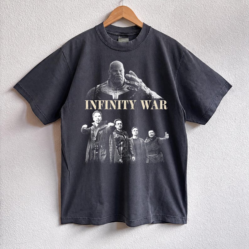 K&ouml;nnte beinhalten: Dunkelgraues T-Shirt mit einer Grafik aus dem Film "Infinity War". Das Design zeigt Charaktere und den Titel in Wei&szlig;. Das T-Shirt h&auml;ngt an einem Holzb&uuml;gel.