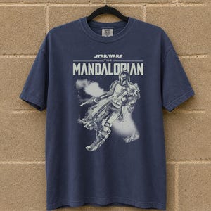 May include: A navy blue t-shirt with the text "STAR WARS THE MANDALORIAN" and a white graphic of the Mandalorian. The graphic shows the character in action with a blaster and smoke effects.