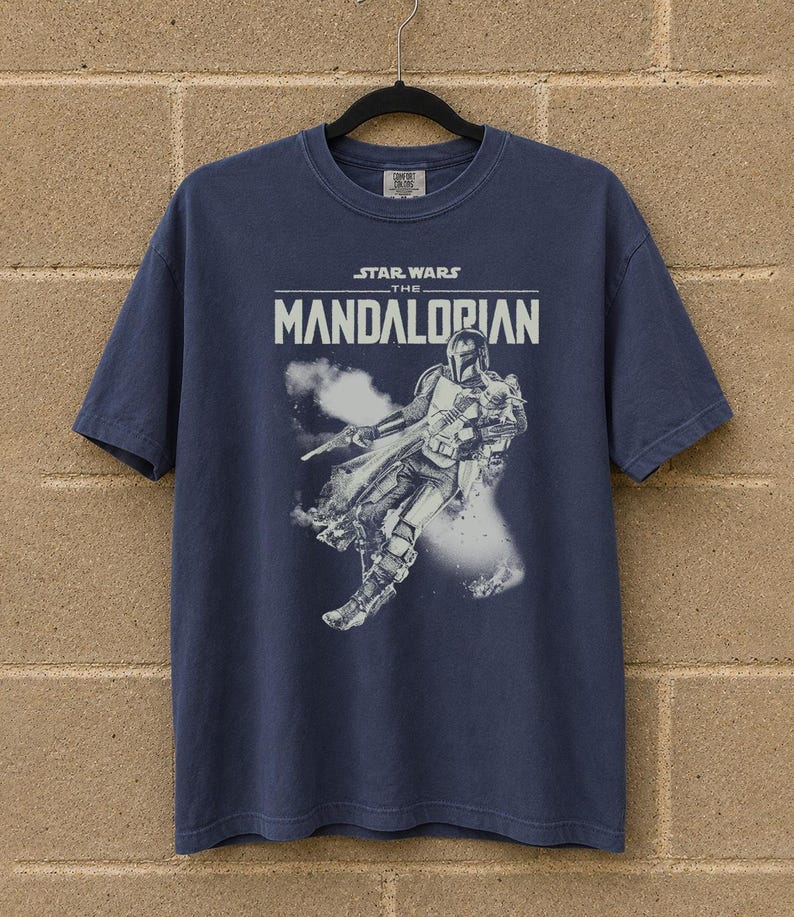 May include: A navy blue t-shirt with the text "STAR WARS THE MANDALORIAN" and a white graphic of the Mandalorian. The graphic shows the character in action with a blaster and smoke effects.