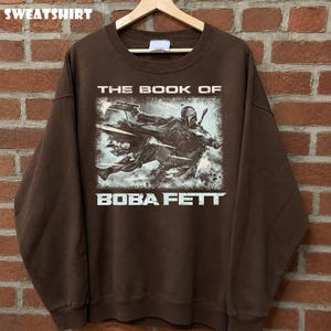 May include: Brown sweatshirt featuring a graphic of Boba Fett from "The Book of Boba Fett." The design shows Boba Fett in action with a blaster. The word "SWEATSHIRT" is printed at the top.
