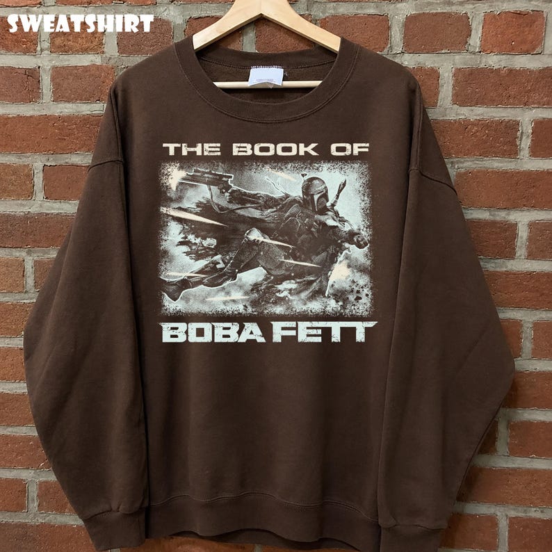 May include: Brown sweatshirt featuring a graphic of Boba Fett from "The Book of Boba Fett." The design shows Boba Fett in action with a blaster. The word "SWEATSHIRT" is printed at the top.