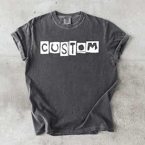 May include: Dark grey t-shirt with rolled-up sleeves. The word "CUSTOM" is printed in white block letters across the front. The shirt has a vintage, worn look.