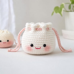 Puede incluir: Una bolsa de ganchillo blanca con cordón que presenta una cara linda con ojos de cuentas negras, una pequeña boca cosida en rosa y mejillas sonrosadas. La bolsa tiene cordones trenzados de color rosa claro y una bola de ganchillo más pequeña.