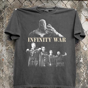 K&ouml;nnte beinhalten: Dunkelgraues T-Shirt mit einer Grafik aus dem Film "Infinity War". Das Design zeigt Thanos und andere Charaktere. Die Worte "INFINITY WAR" sind in Beige &uuml;ber die Mitte des T-Shirts gedruckt.