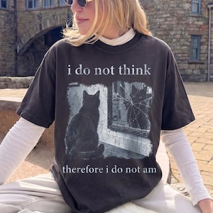 May include: A charcoal gray short-sleeve t-shirt featuring a graphic of a black cat sitting before a cracked mirror. The text "i do not think" appears above the cat, and "therefore i do not am" is printed below the graphic in a white, distressed font.