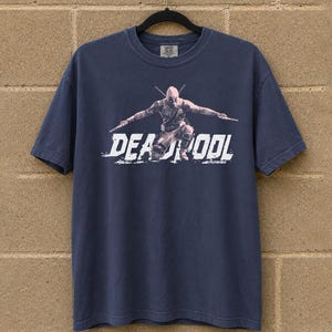 May include: Navy blue t-shirt with a graphic of Deadpool and the word "DEADPOOL" in white, distressed lettering. The superhero is shown in a dynamic pose, holding two swords. The shirt is hanging on a black hanger against a brick wall.