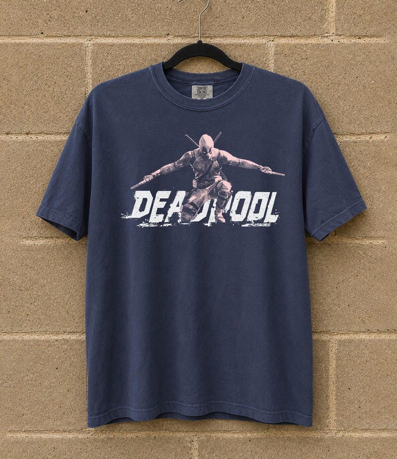 May include: Navy blue t-shirt with a graphic of Deadpool and the word "DEADPOOL" in white, distressed lettering. The superhero is shown in a dynamic pose, holding two swords. The shirt is hanging on a black hanger against a brick wall.