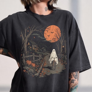 May include: A charcoal gray t-shirt featuring a graphic print of a ghost wearing a white sheet and riding a bicycle down a path at night. The scene includes a large orange moon, bare trees, a house, pumpkins, and bats in a dark, autumnal setting.