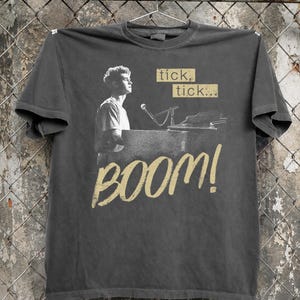 May include: Dark gray t-shirt featuring a black and white image of a person at a piano with a microphone. The shirt has the words "tick, tick..." and "BOOM!" in a gold color.