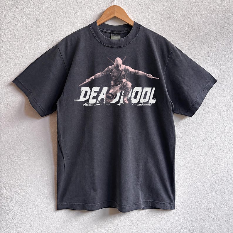 May include: Dark grey t-shirt with a Deadpool graphic, crossed katanas, and the word "DEADPOOL" in distressed white lettering. The tee is suspended on a wooden hanger against a white wall.