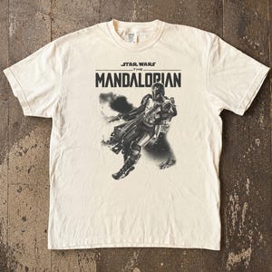 May include: Cream-coloured t-shirt with a graphic of The Mandalorian from Star Wars. The design includes the text "STAR WARS THE MANDALORIAN" above a black and white illustration of the character. The shirt has a crew neck and short sleeves.