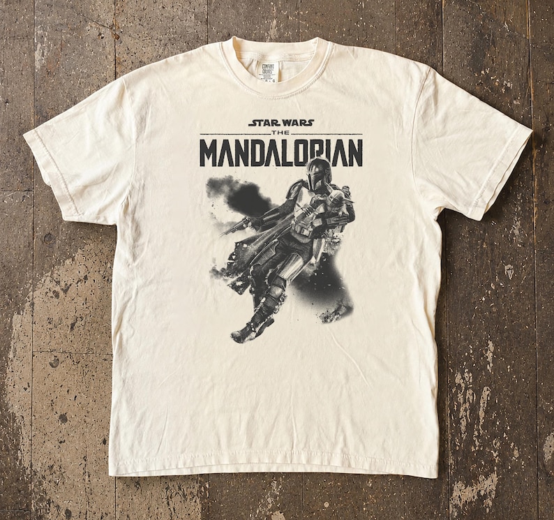 May include: Cream-coloured t-shirt with a graphic of The Mandalorian from Star Wars. The design includes the text "STAR WARS THE MANDALORIAN" above a black and white illustration of the character. The shirt has a crew neck and short sleeves.