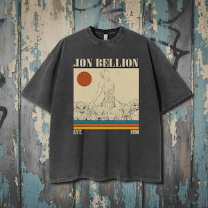 Può includere: T-shirt grigio scuro con un design grafico. Il design include il testo "JON BELLION", un'illustrazione di una figura e pecore, un sole rosso e linee a strisce. Il testo "EST. 1990" è sotto.