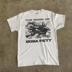 May include: White t-shirt featuring a graphic of Boba Fett from "The Book of Boba Fett". The image is in black and white, with the text "THE BOOK OF" above and "BOBA FETT" below the graphic. The shirt has a crew neck.