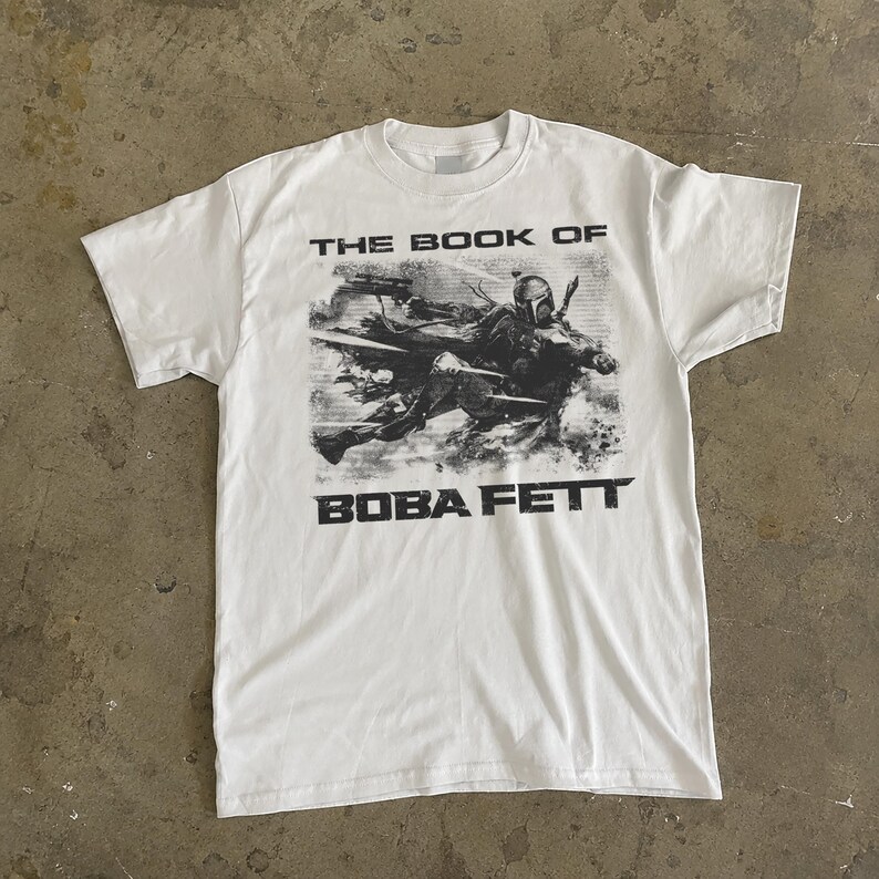 May include: White t-shirt featuring a graphic of Boba Fett from "The Book of Boba Fett". The image is in black and white, with the text "THE BOOK OF" above and "BOBA FETT" below the graphic. The shirt has a crew neck.