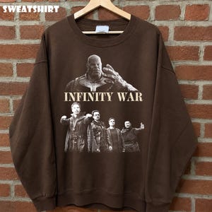 K&ouml;nnte beinhalten: Braunes Sweatshirt mit einer Grafik aus dem Film "Infinity War". Das Design enth&auml;lt den Text "INFINITY WAR" und Bilder von Charakteren. Das Wort "SWEATSHIRT" ist oben aufgedruckt.