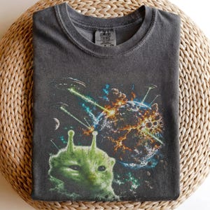 May include: A charcoal grey crew neck t-shirt featuring a graphic print of a green alien head in the foreground. The background depicts a planet exploding in space with multiple flying saucers firing green laser beams toward the planet surface.