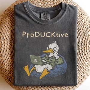 以下が含まれることがあります： チャコールグレーの半袖Tシャツ。緑色のパーカーを着た白いアヒルが青いビーズクッションに座り、ノートパソコンを使っているグラフィックが描かれています。アヒルの上には、かすれたベージュ色のフォントで「ProDUCKtive」という文字がプリントされています。