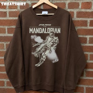 May include: A brown sweatshirt with a graphic of The Mandalorian from Star Wars. The design includes the text "STAR WARS THE MANDALORIAN" in white. The sweatshirt is displayed on a wooden hanger.