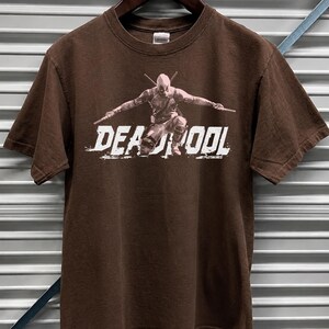 May include: Dark chocolate brown t-shirt with a Deadpool graphic and the word "DEADPOOL" in white. The t-shirt is displayed on a wooden hanger against a corrugated metal background.